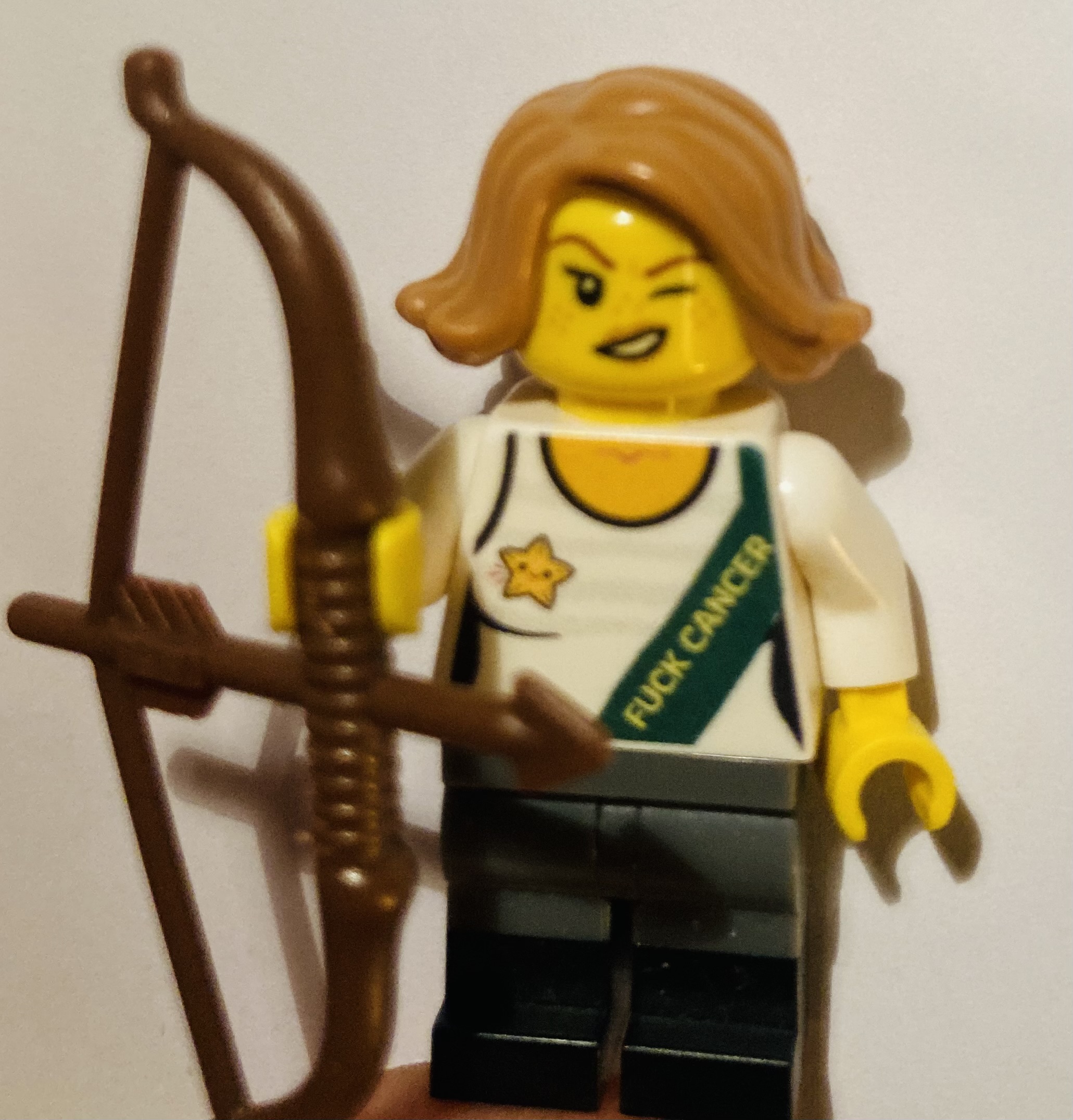 Eine zwinkernde Lego-Figur mit blondem Haar hält Pfeil und Bogen in der Hand. Sie trägt ein Top mit der Aufschrift "Fuck Cancer"